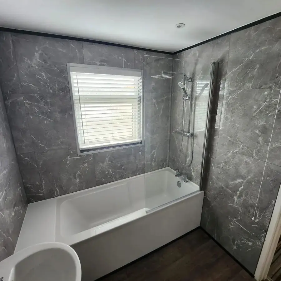 A bathroom with grey marble effect wall panels and a bath with shower over