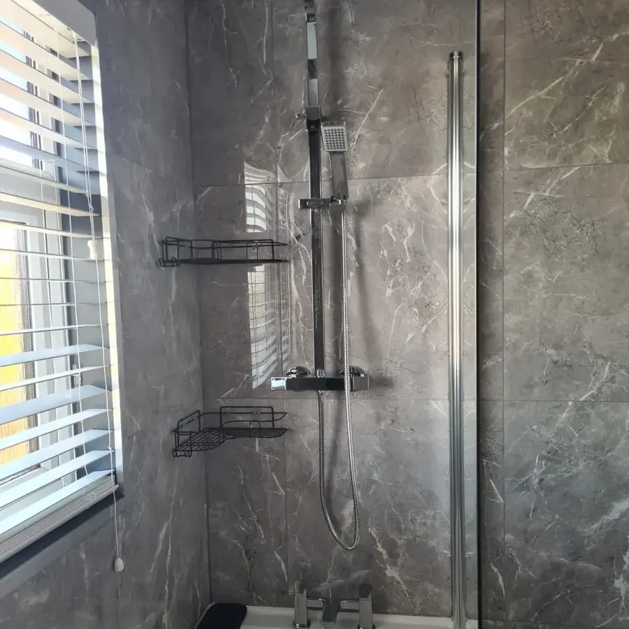 A shower over a bath with grey marble effect panelling and a glass screen